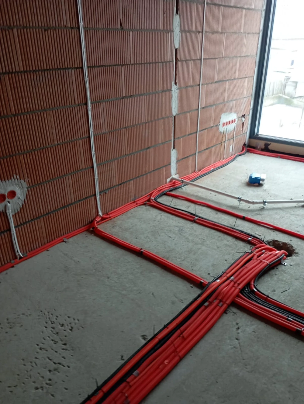 Ułożone na surowej betonowej posadzce czerwone i czarne peszle z przewodami przy murowanej ścianie – instalacje elektryczne