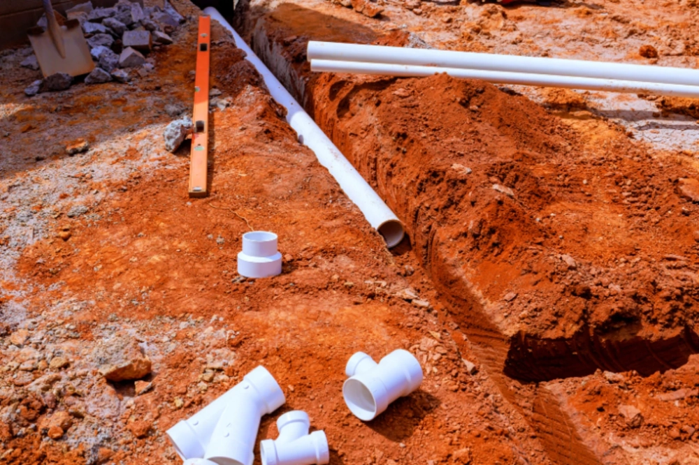 Białe rury i kształtki PVC w wykopanym rowie na czerwonej ziemi – instalacje hydrauliczne